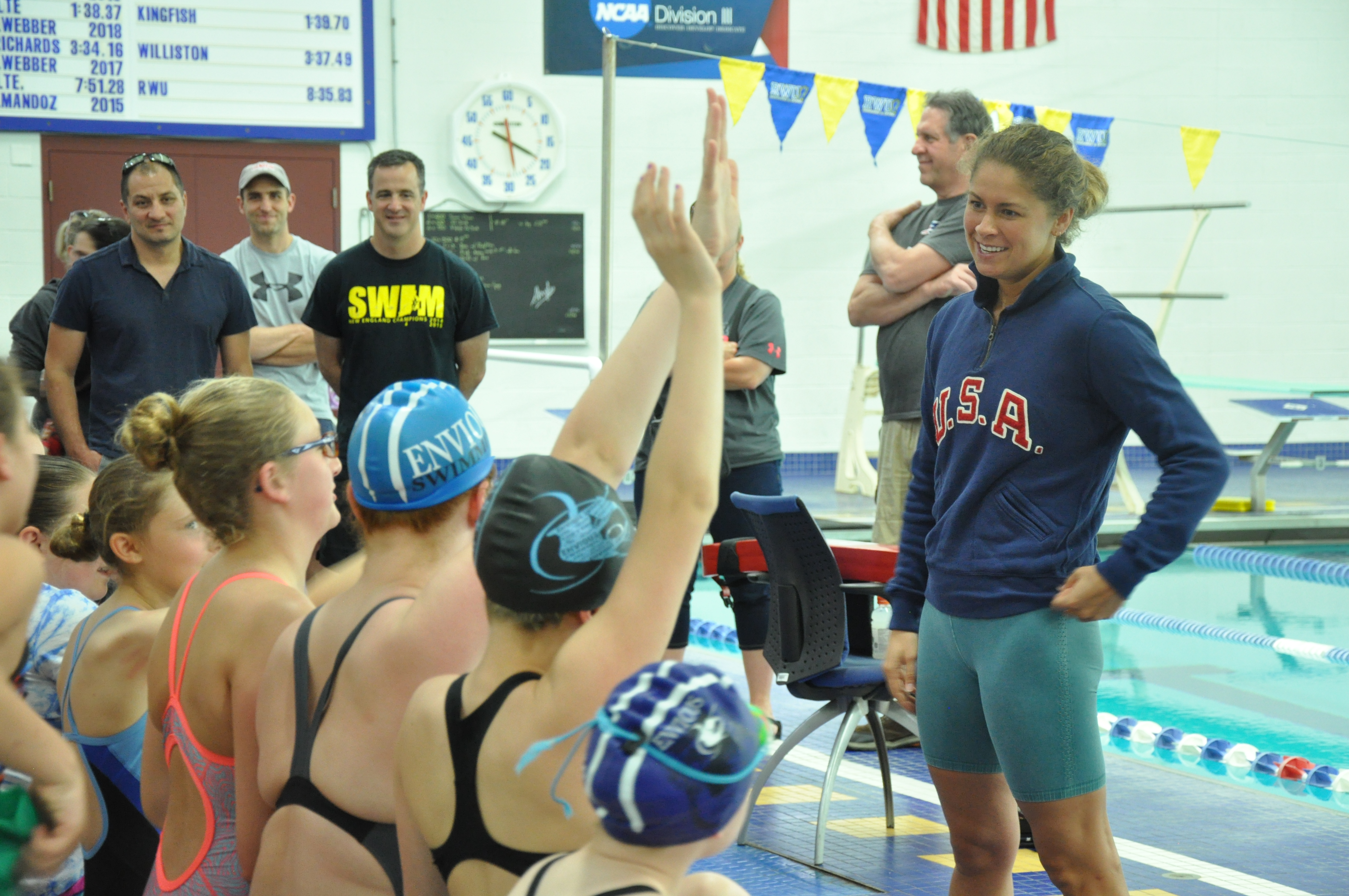 A group of young swimmers face away from the camera, raising hands. Olympic swimmer Elizabeth Beisel stands smiling before them, taking questions, wearing a blue 